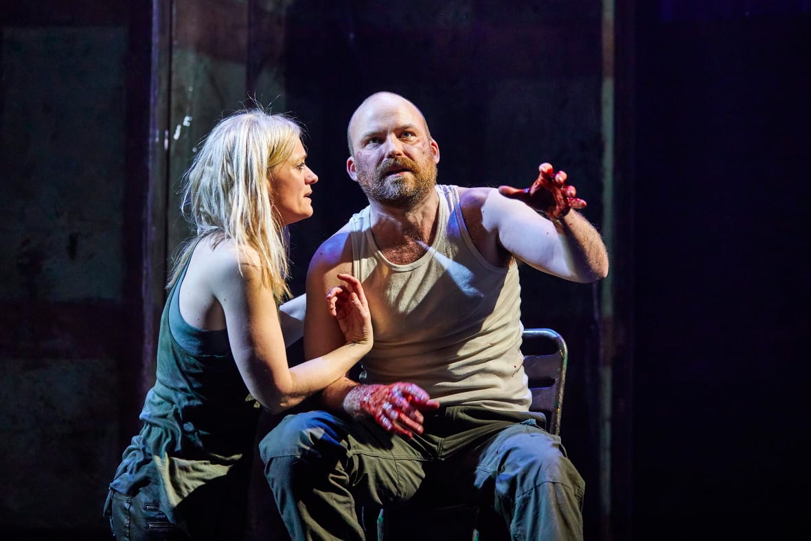 A man sits in a chair on stage during a production of Macbeth. His hands are covered in blood and he stared ahead and reaches out with one hand towards the camera. A woman crouches next to him with her arm around his shoulder looking at his face. Credit line: Macbeth 2018 photo by Brinkhoff Mögenburg